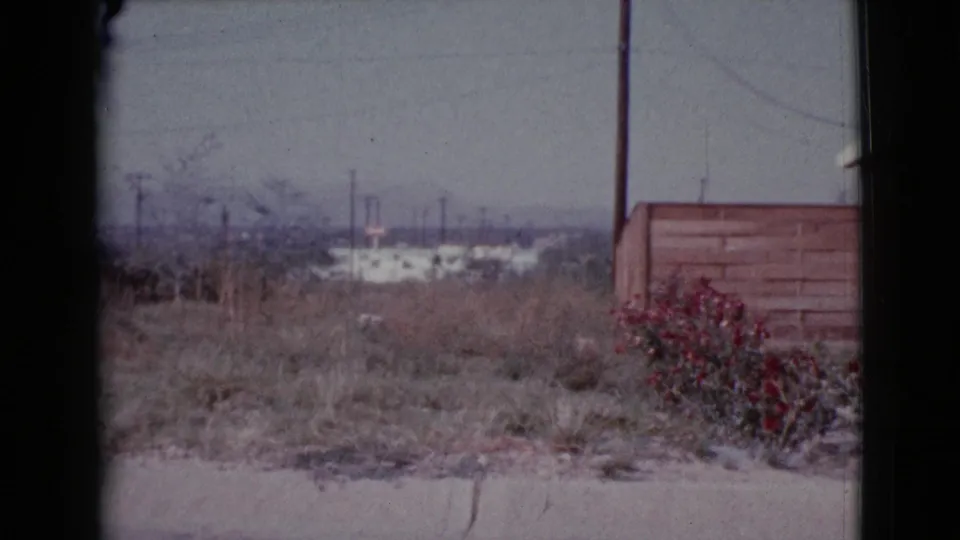 1964 Cottonwood, Arizona frame showing a field edge with shrubs, a fence, utility pole, and distant town structures.