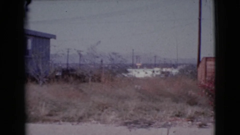 Cottonwood, Arizona home-movie frame from 1964 with a small shed or trailer at the field edge and distant town buildings beyond.