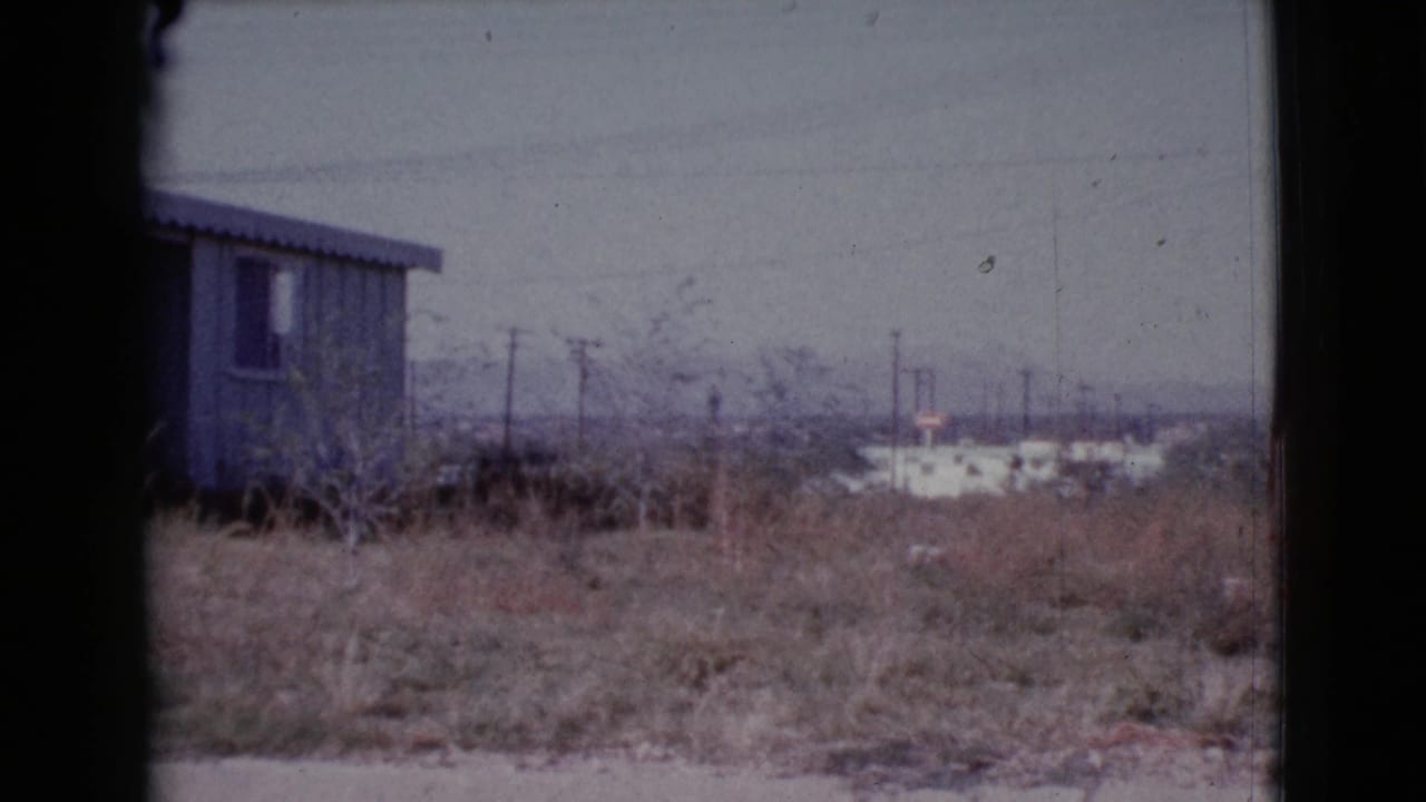 Small weathered house beyond dry grass, seen through a dark window or frame.
