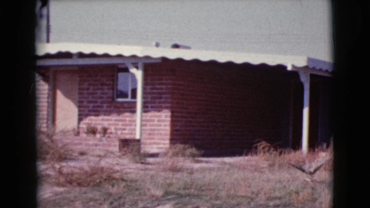 Front portion of a brick house with porch posts, small window, dry yard, and bright sky.