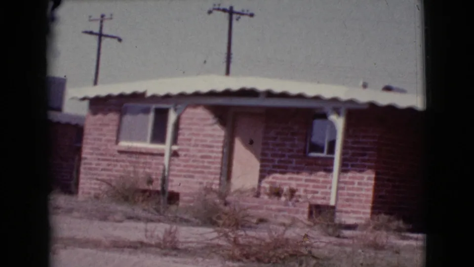 A slightly shifted 1964 home-movie frame shows the brick house facade, porch awning, windows, dry vegetation, and poles in Cottonwood, Arizona.