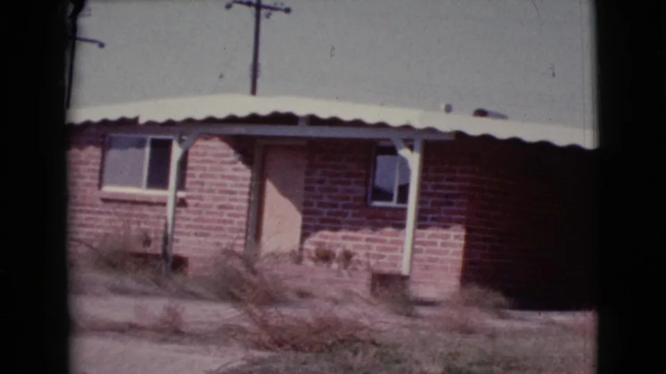 Final frame of the 1964 Cottonwood house exterior, showing the small brick residence, covered porch, dry yard plants, curb, and utility poles.