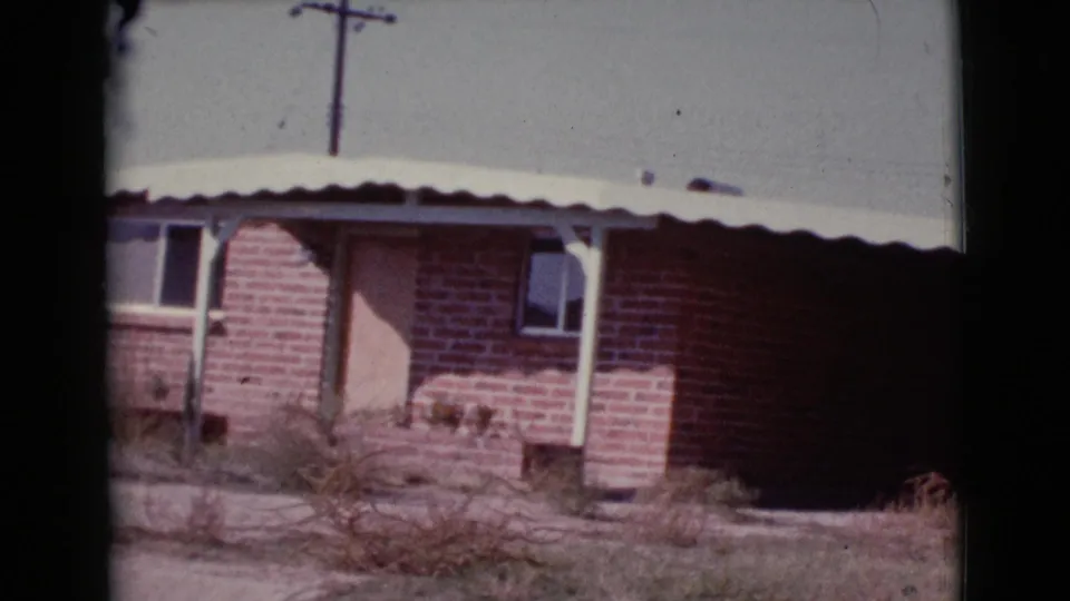 1964 Cottonwood, Arizona 8mm frame showing a red brick house with a light door, small windows, white awning, dry yard, and utility pole.