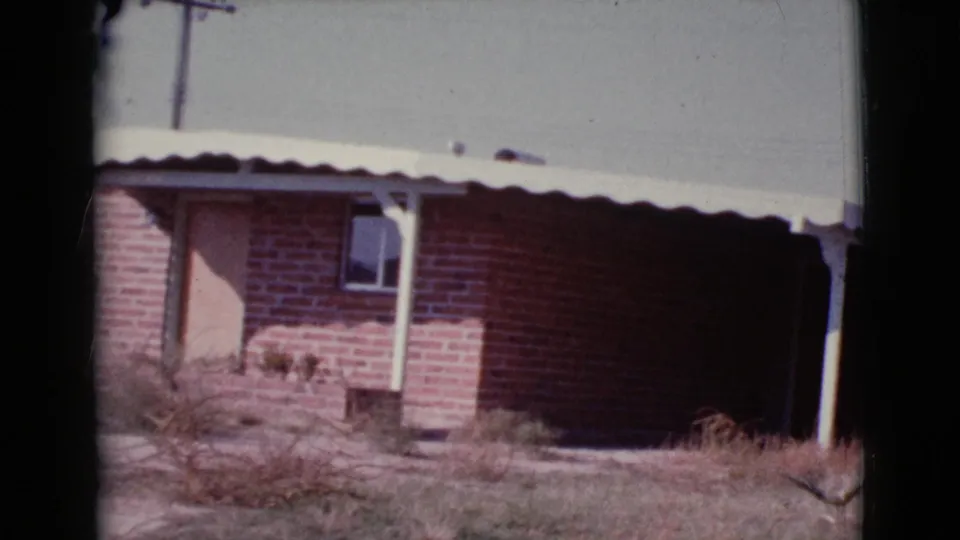 Cottonwood, Arizona house exterior on 8mm film, with red brick walls, a white roof edge, a light door, windows, dry grass, and a utility pole.