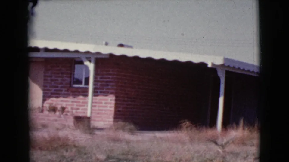 1964 8mm frame of the red brick house from a slightly shifted angle, showing the awning, a window, shaded wall, and dry vegetation.