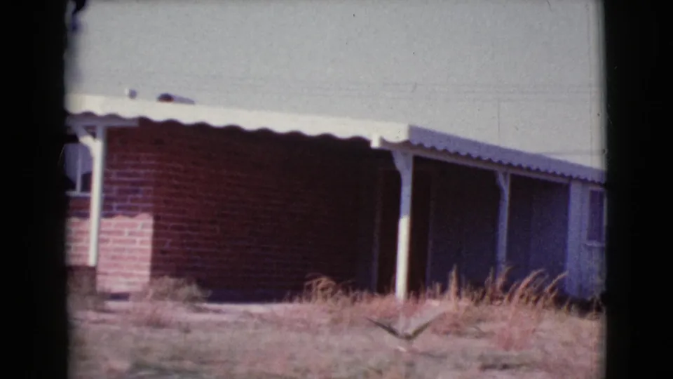 Cottonwood, Arizona 8mm frame showing the brick house side, white awning, support posts, dry grass, and the edge of an adjoining structure.