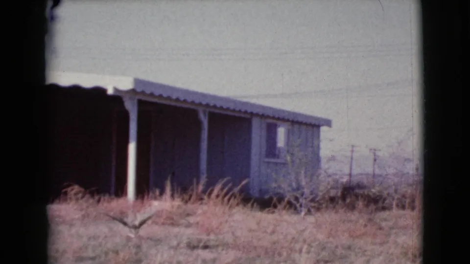 1964 Cottonwood, Arizona frame of a small house exterior with brick, blue siding, shaded porch, dry grass, and utility poles in the distance.
