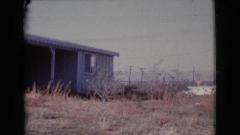 1964 Cottonwood, Arizona home-movie frame as the pan moves past the blue-sided house toward dry brush, poles, and distant low buildings.