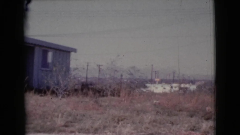 Final 1964 Cottonwood, Arizona frame showing the house partly cropped at left, with dry vegetation, utility poles, and low distant structures.