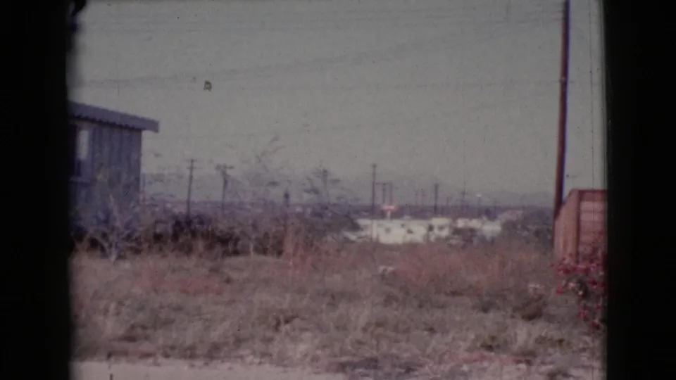 1964 Cottonwood, Arizona home-movie frame showing dry ground, utility poles, distant buildings, a wooden shed, and red flowers.