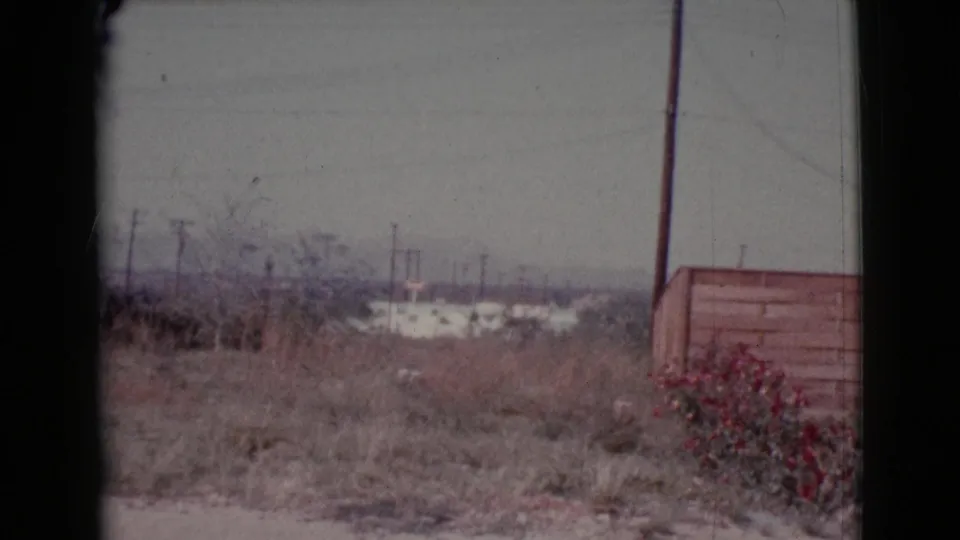 1964 Cottonwood, Arizona view across a dry yard toward utility poles, low distant buildings, a fence, and red flowers.