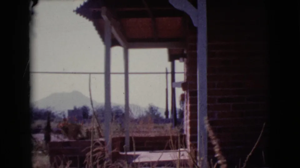 1964 Cottonwood, Arizona home-movie frame from a porch, with brickwork, posts, dry vegetation, and hazy hills beyond.