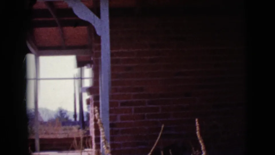 1964 Cottonwood, Arizona frame of a red brick exterior porch with white posts, sunlit opening, dry vegetation, and distant hills.