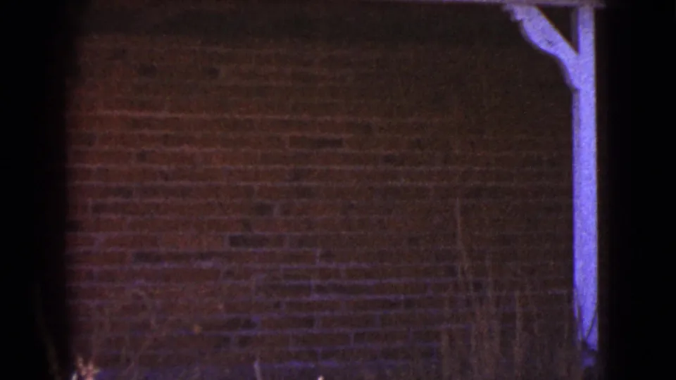 1964 Cottonwood, Arizona 8mm frame showing a dim brick house wall and white-painted porch beam in low light.