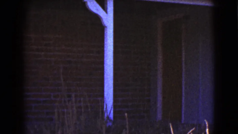 Low-light 8mm view of a brick house exterior in Cottonwood, with a painted support post and sparse plants near the wall.