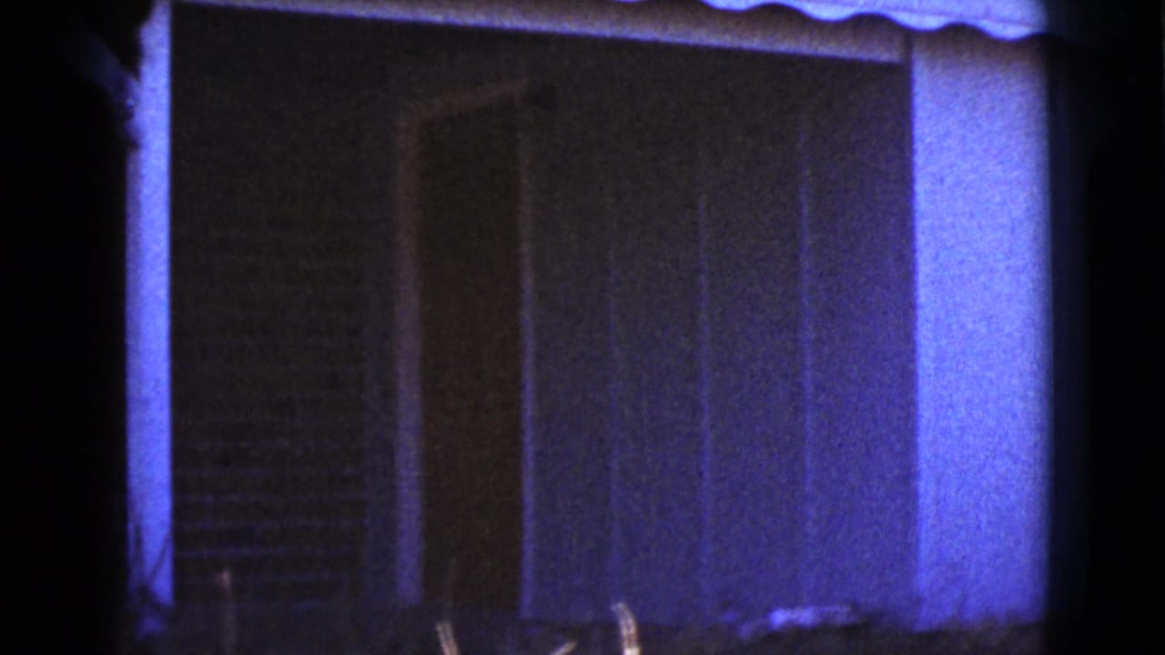 Blue-tinted 8mm frame of a shaded house wall with vertical siding and a dark window area.