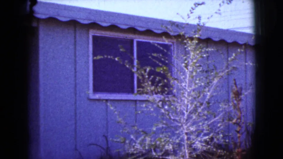 1964 Cottonwood, Arizona frame showing the house window and siding with sunlit brush as the pan reaches the right side.