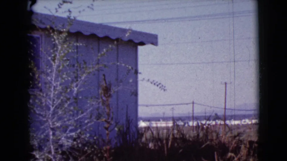 As the camera pans right, the house edge gives way to open Cottonwood landscape with power lines, poles, brush, and distant low structures.
