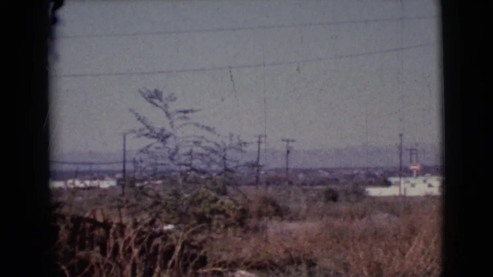 1964 8mm rural Cottonwood frame with overgrown grasses, shrubs, utility wires, distant white buildings, and a small vertical structure.