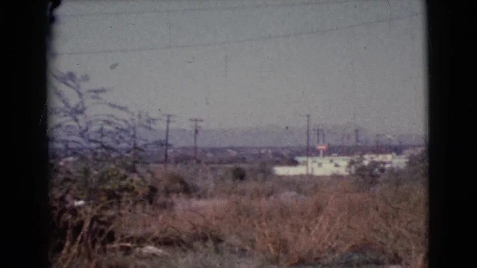 Cottonwood, Arizona home-movie frame showing dry roadside vegetation, power lines, a distant white building, and hazy hills.