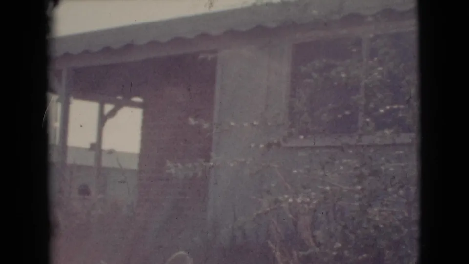 1964 8mm view of a modest house exterior in Cottonwood, Arizona, with porch posts, roof edge, and branches in front.