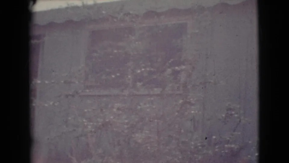 Faded 1964 8mm frame showing a Cottonwood house exterior with roofline, wall, window area, and branches crossing the view.