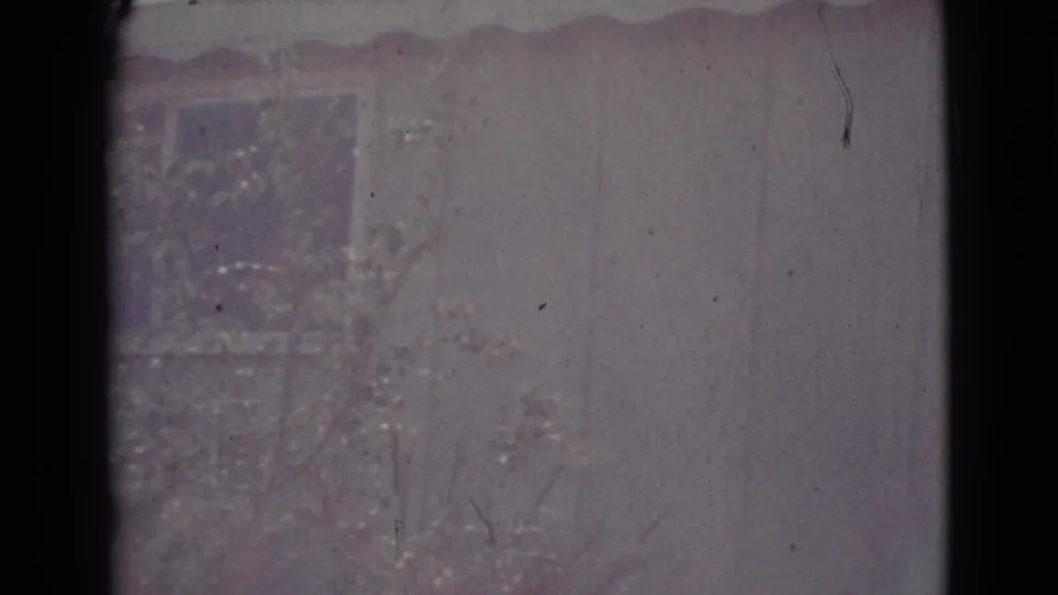 1964 Cottonwood home-movie frame with a pale residential wall, roof edge, and foreground vegetation softened by faded 8mm color.