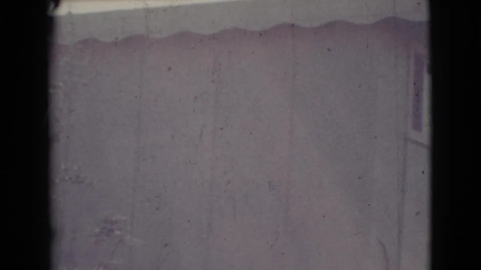 Late frame of the 1964 Cottonwood house exterior, showing a pale wall, corrugated roof edge, and faint vegetation through faded 8mm exposure.