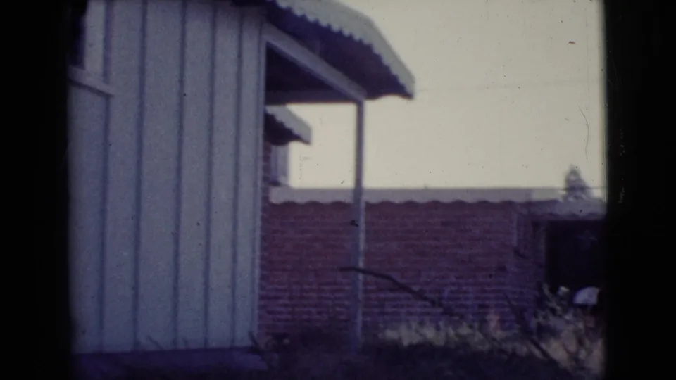 1964 Cottonwood, Arizona frame showing a house edge, porch post, low brick structure, and dry yard vegetation in soft 8mm color.