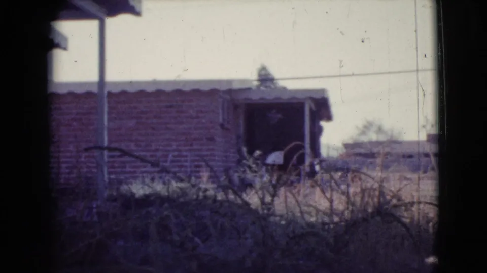 1964 Cottonwood, Arizona 8mm frame showing a brick structure with a dark opening, roof edge, porch post, and overgrown dry vegetation.