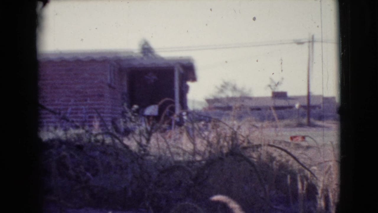Dry field with brush, a low brick building, distant structures, and overhead utility wires.