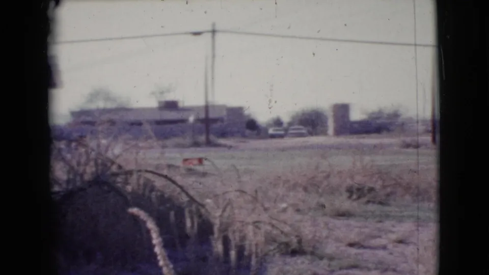 1964 Cottonwood, Arizona frame with open dry ground, brush, low settlement buildings, a utility pole, overhead wires, and a small distant vehicle-like shape.