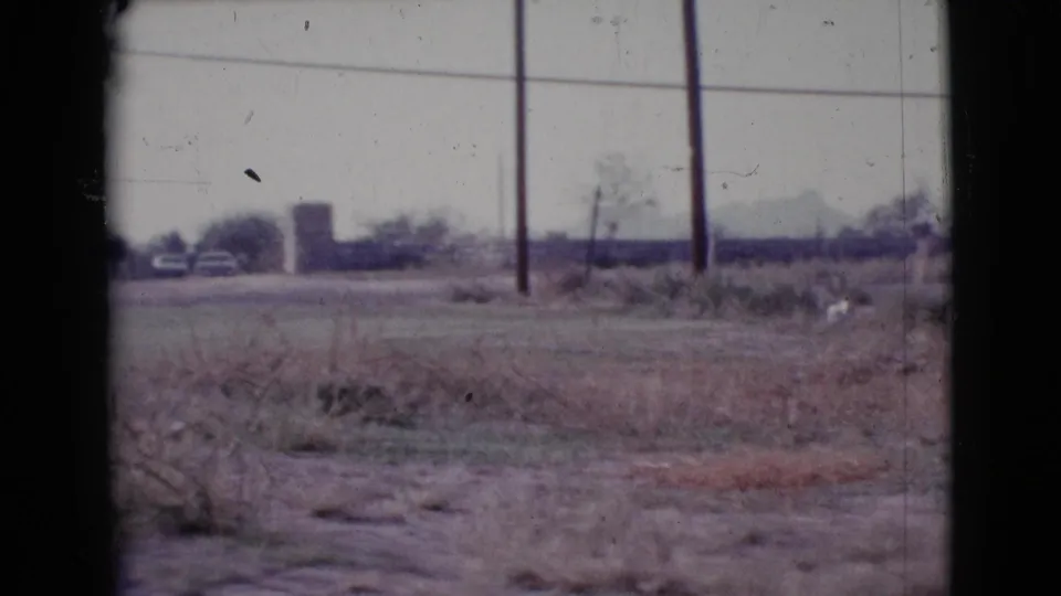 1964 Cottonwood, Arizona frame showing dry open land, sparse brush, two utility poles, overhead lines, and distant low buildings.