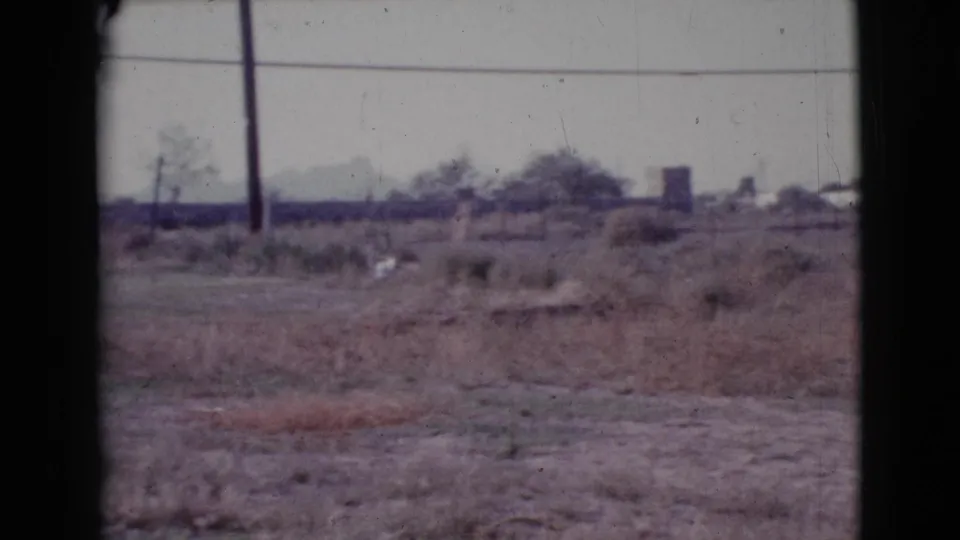1964 Cottonwood, Arizona home-movie frame with open dry land, utility poles, power lines, brush, and low distant structures on the horizon.