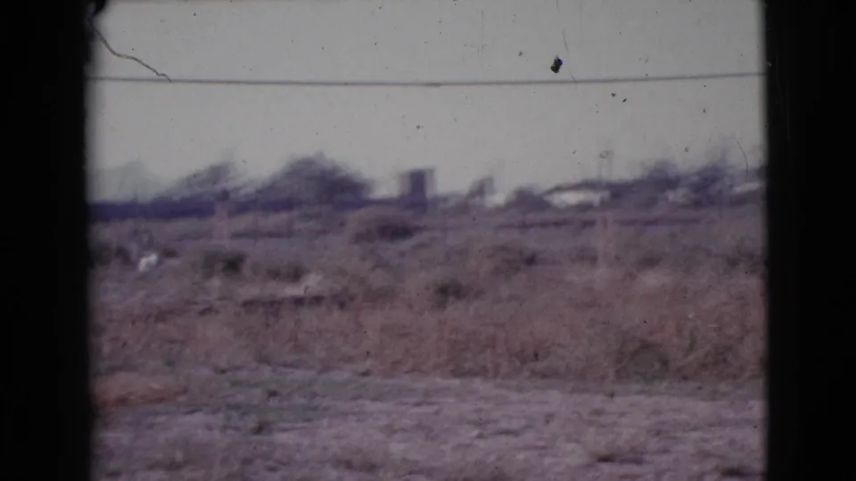 1964 Cottonwood, Arizona 8mm frame showing dry outskirts with scrub, distant low structures, utility wires, and a pale sky.