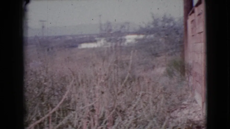 Faded 8mm frame from Cottonwood, Arizona showing dry plants in the foreground and pale distant rooftops or structures.