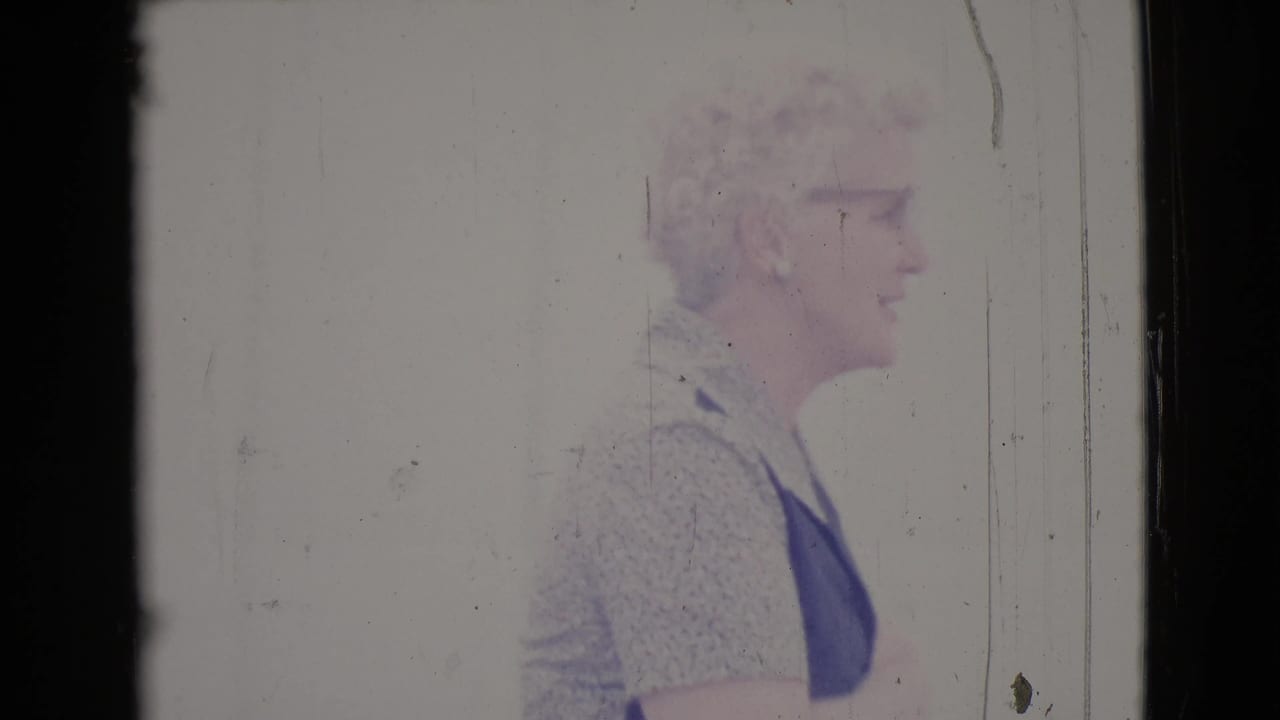 White-haired woman in glasses seen in profile outside a pale building.
