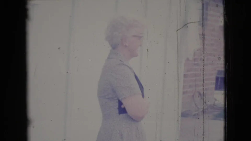 A 1964 home-movie frame shows the woman standing near a pale wall, with brickwork visible at the side.