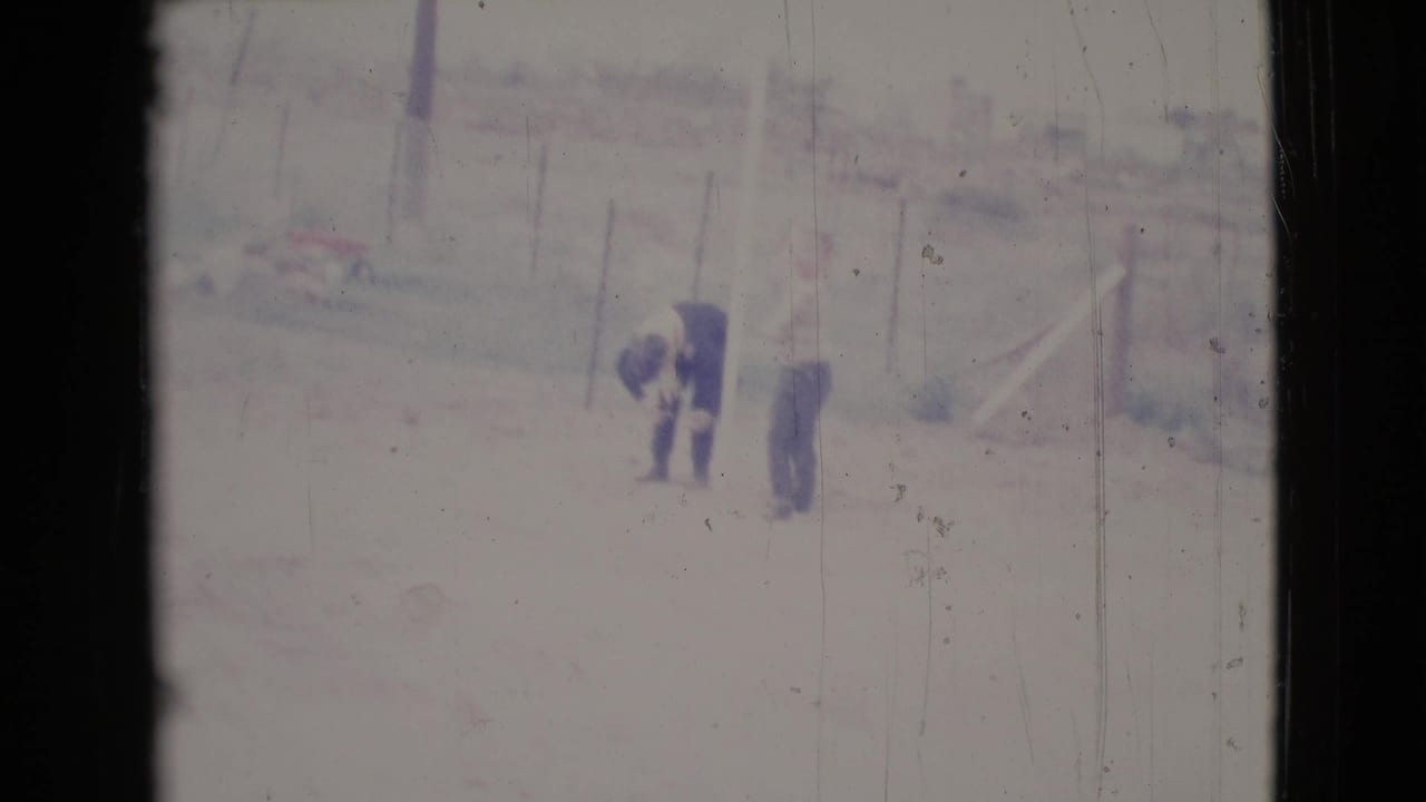 In 1964 Cottonwood, Arizona footage, two distant adults stand in a dusty fenced yard, with one bending slightly beside the other.