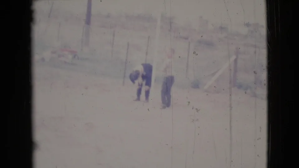 In 1964 Cottonwood, Arizona footage, two distant adults stand in a dusty fenced yard, with one bending slightly beside the other.