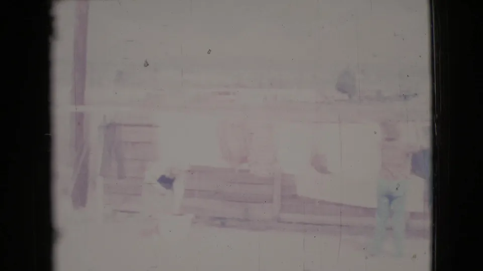 Two women are faintly visible in a 1964 home-movie frame as they work around a clothesline near a porch railing in Cottonwood.
