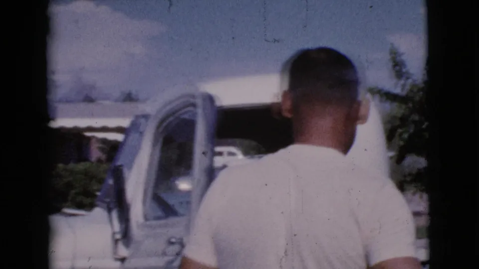 1964 Cottonwood, Arizona frame showing a man from behind as he stands near an open vehicle door in daylight.