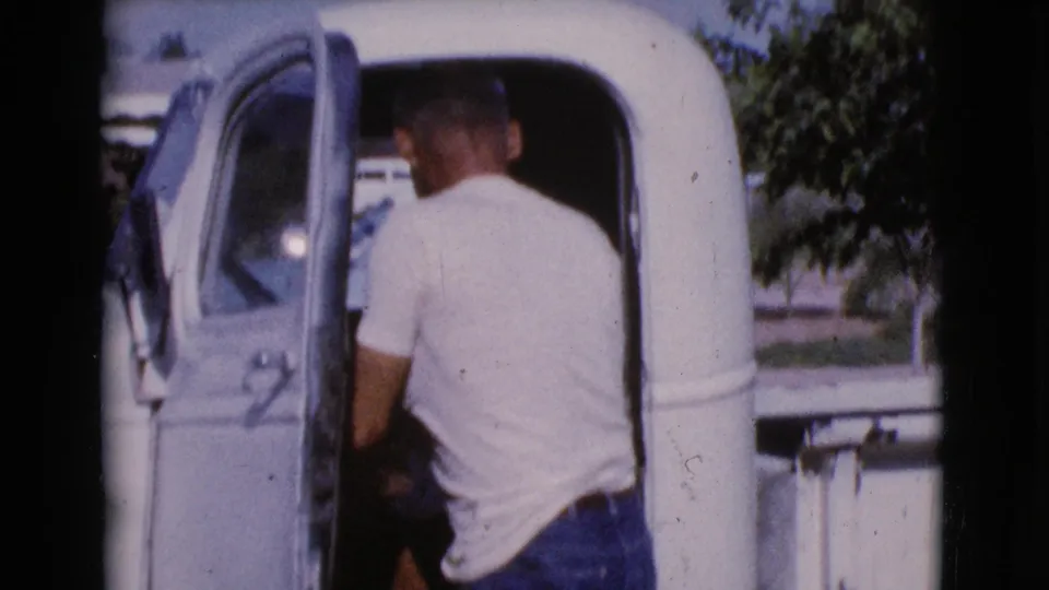 The man pauses at the open driver's side of a white pickup truck, seen from behind in sunlit 1964 Cottonwood home-movie footage.