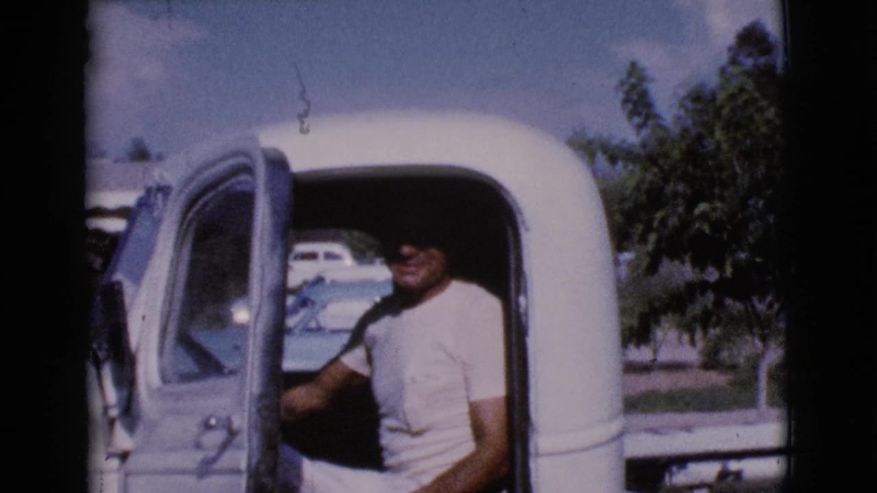 Man in a white T-shirt seated in the open doorway of a vintage pickup truck.