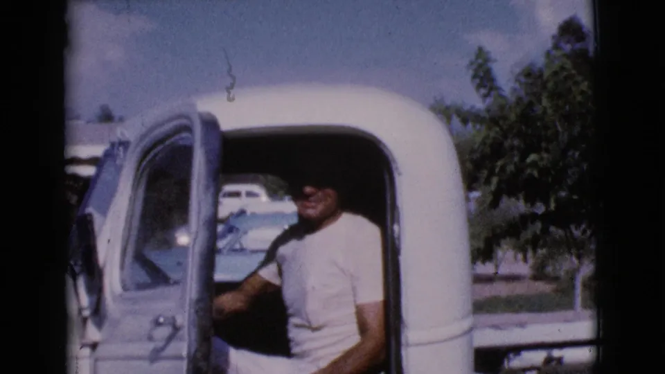 In 1964 Cottonwood, Arizona footage, a man sits in the driver's seat of a vintage pickup with the door open on a sunny residential street.
