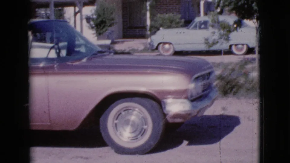 A two-tone purple and white vintage sedan is parked along a sunny Cottonwood, Arizona residential street in 1964 home-movie footage.
