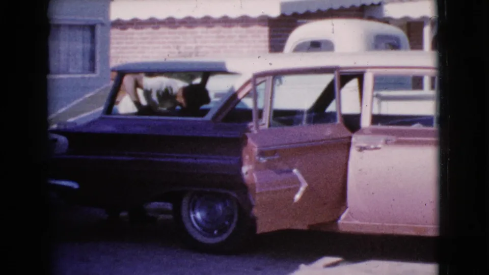 1964 Cottonwood, Arizona home-movie frame showing a man partly visible as he works near the back of a two-tone station wagon.