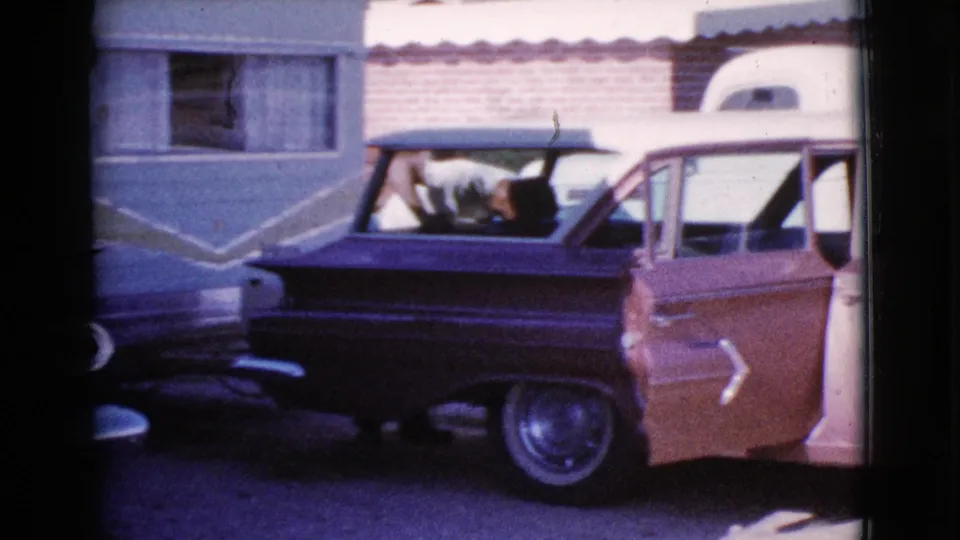 1964 Cottonwood, Arizona frame of a two-tone station wagon and white RV trailer as a man leans into the vehicle during travel preparation.