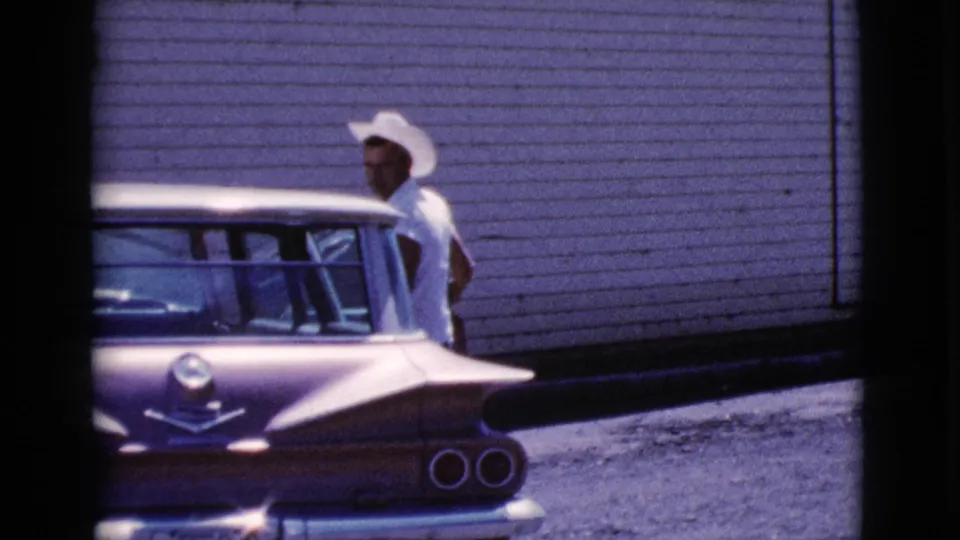 In the 1964 Cottonwood home movie, a man in a white cowboy hat stands next to a purple and white sedan.