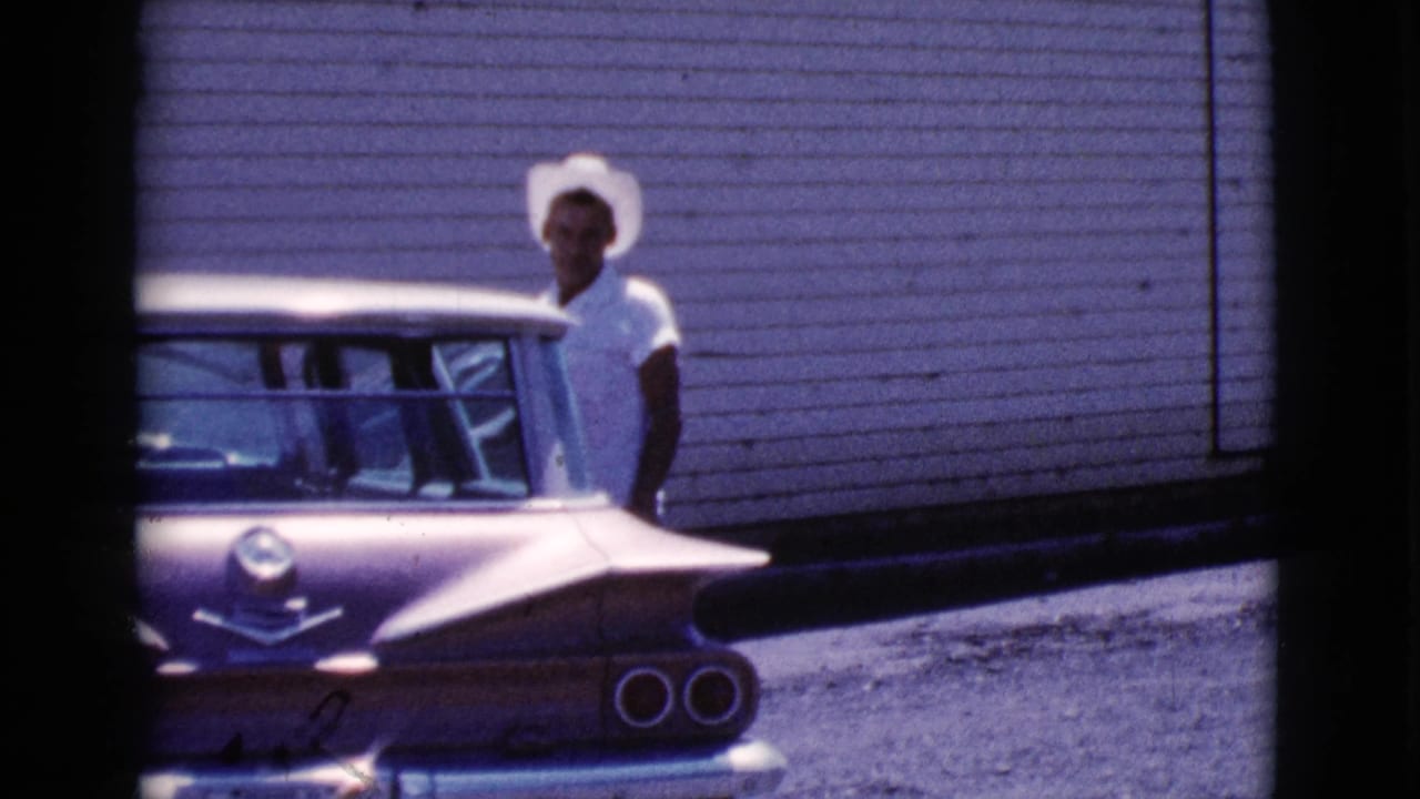Young man in a white cowboy hat stands beside the rear of a tailfin-era car.